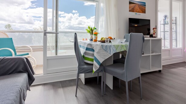 A modern dining area with a table set for two, a television, and a view of the sky through large windows.