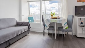 A modern living room with a grey sofa, a dining table with two chairs, a TV on a stand, and a large window with a view of the sky and clouds.