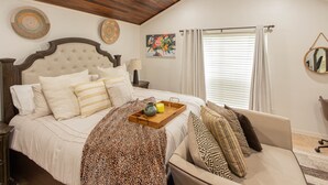 Een slaapkamer met een groot bed, een houten hoofdeinde, een nachtkastje met een lamp, een raam met jaloezieën en een schilderij aan de muur.