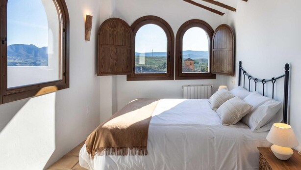 A bedroom with a large bed, wooden beams, and three arched windows offering a view of the landscape. - Magnificent rustic house with pool, garden, and views (Acequias)