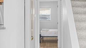 Un couloir étroit mène à une chambre à coucher qui contient un lit, une fenêtre avec des stores et un plancher en bois.