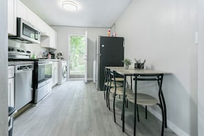 Kompakti keittiö, jossa on ruostumattomasta teräksestä valmistetut kodinkoneet, ruokailutila pöydän ja tuolien kera sekä näkymä vehreään maisemaan.