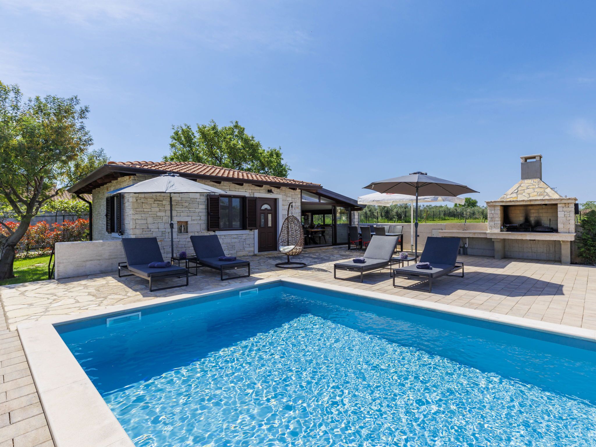 Une piscine avec des chaises longues, un bâtiment en pierre doté d’un toit en tuiles et une cheminée en pierre.