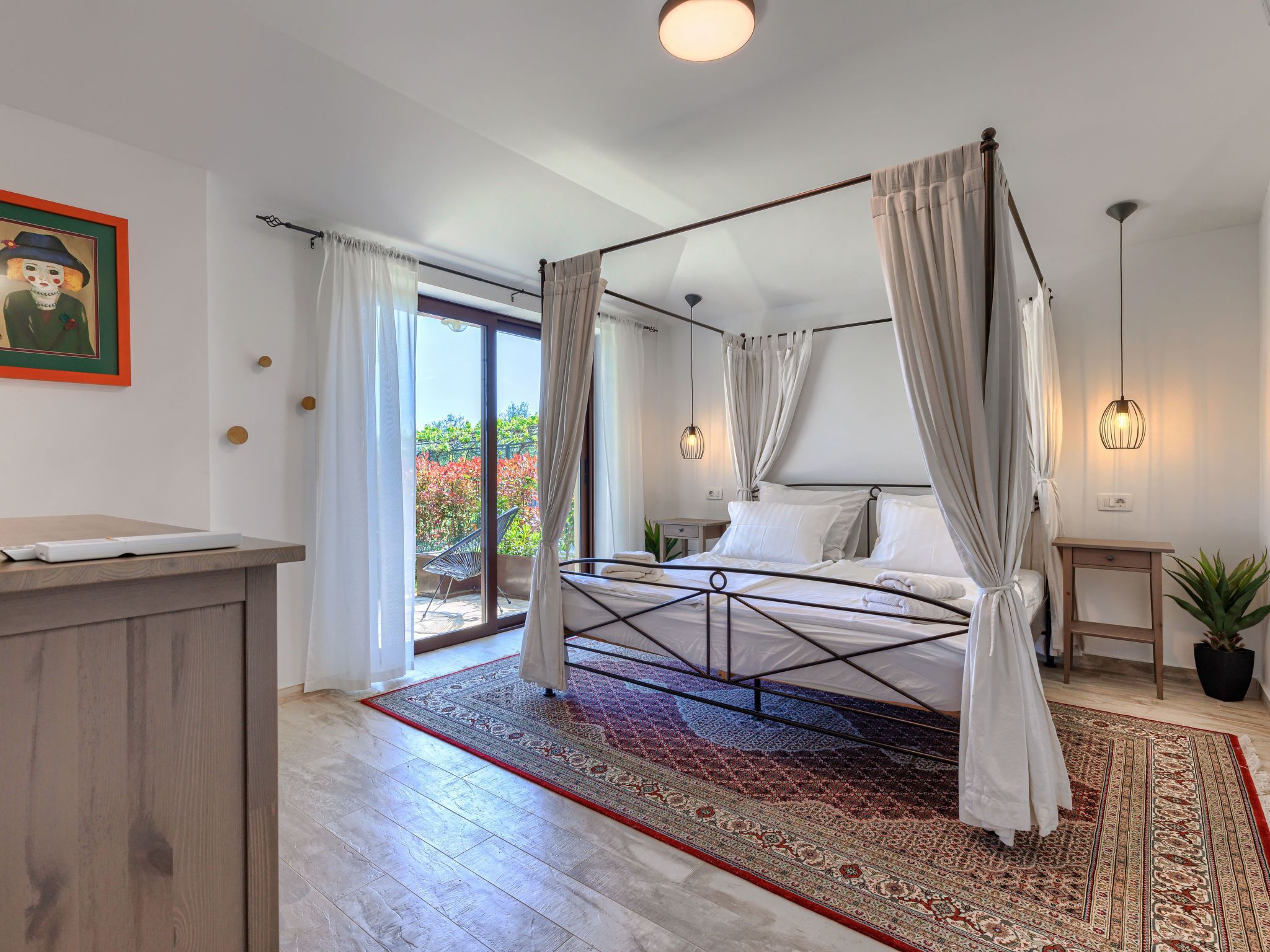 A spacious bedroom with a four-poster bed, a large window with curtains, a wooden desk, and a potted plant.