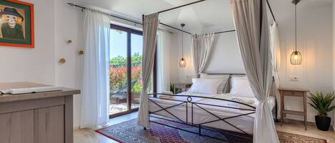 A spacious bedroom with a four-poster bed, a large window with curtains, a wooden desk, and a potted plant.