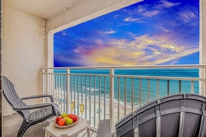 A balcony with a view of the ocean, a table with fruit and drinks, and two chairs.