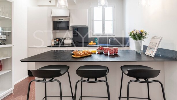 Una cucina moderna con isola centrale, sgabelli da bar neri, illuminazione a sospensione e vista sull'area pranzo.