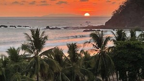 Un atardecer sobre una zona costera con palmeras y afloramientos rocosos.