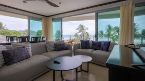 Una sala moderna con un sofá, dos mesas de centro y una televisión. Amplias ventanas ofrecen vistas a palmeras y al océano.