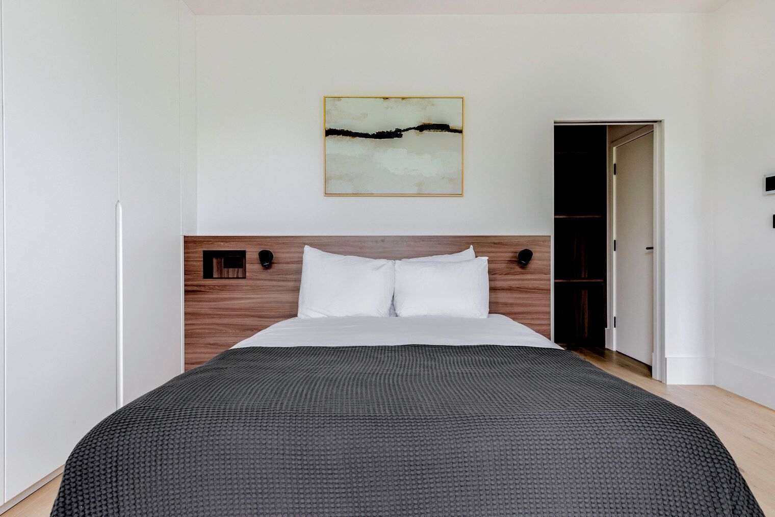 A modern bedroom with a wooden headboard, a large bed with white bedding, a textured throw blanket, and a minimalist painting above the bed.