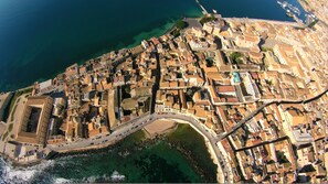 Vista aérea de uma cidade costeira com mistura de edifícios históricos e modernos, uma estrada curva proeminente e uma visão clara do mar adjacente.