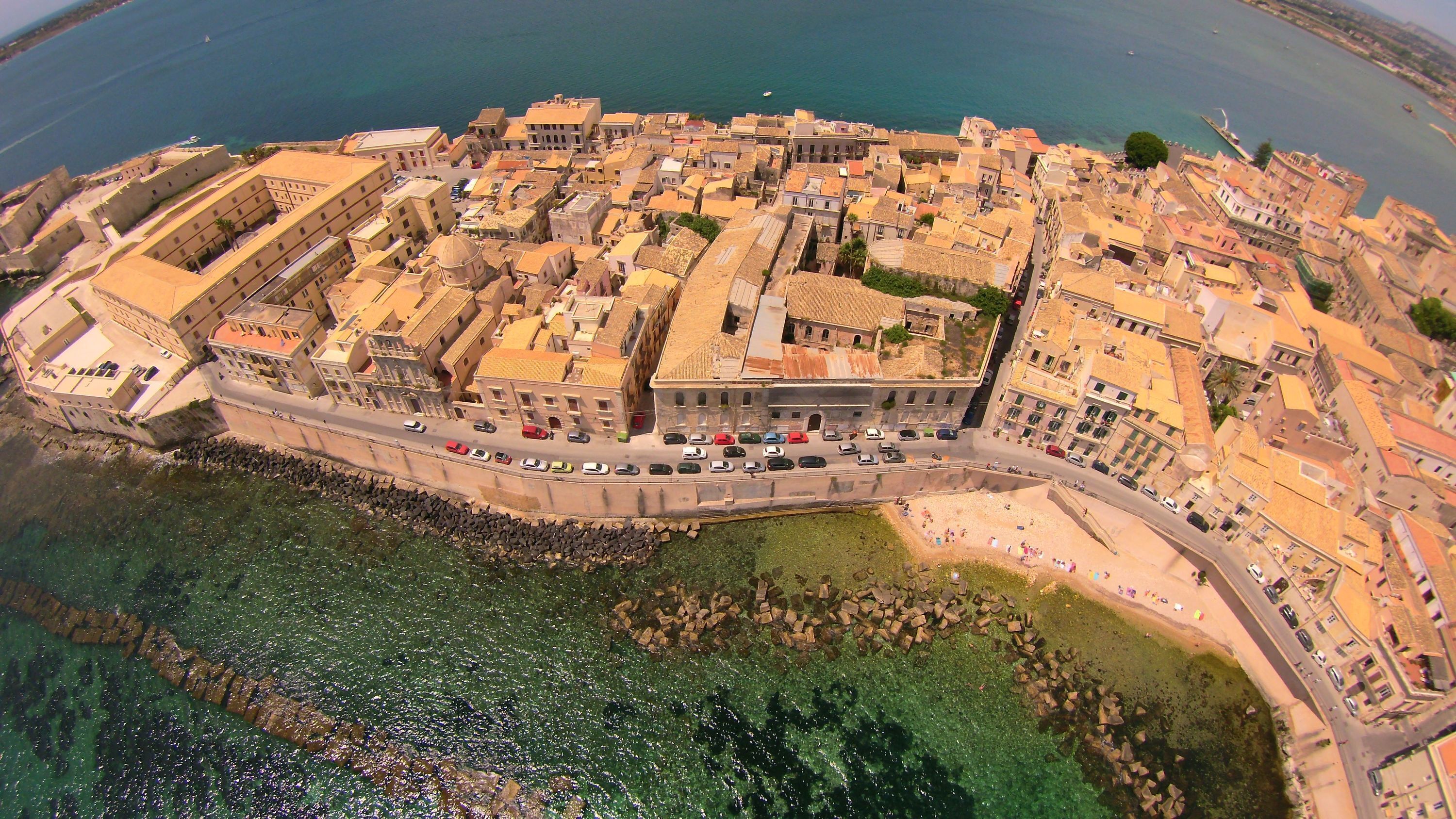 An aerial view of a coastal town with historic buildings, a fortified structure, and a clear view of the sea.