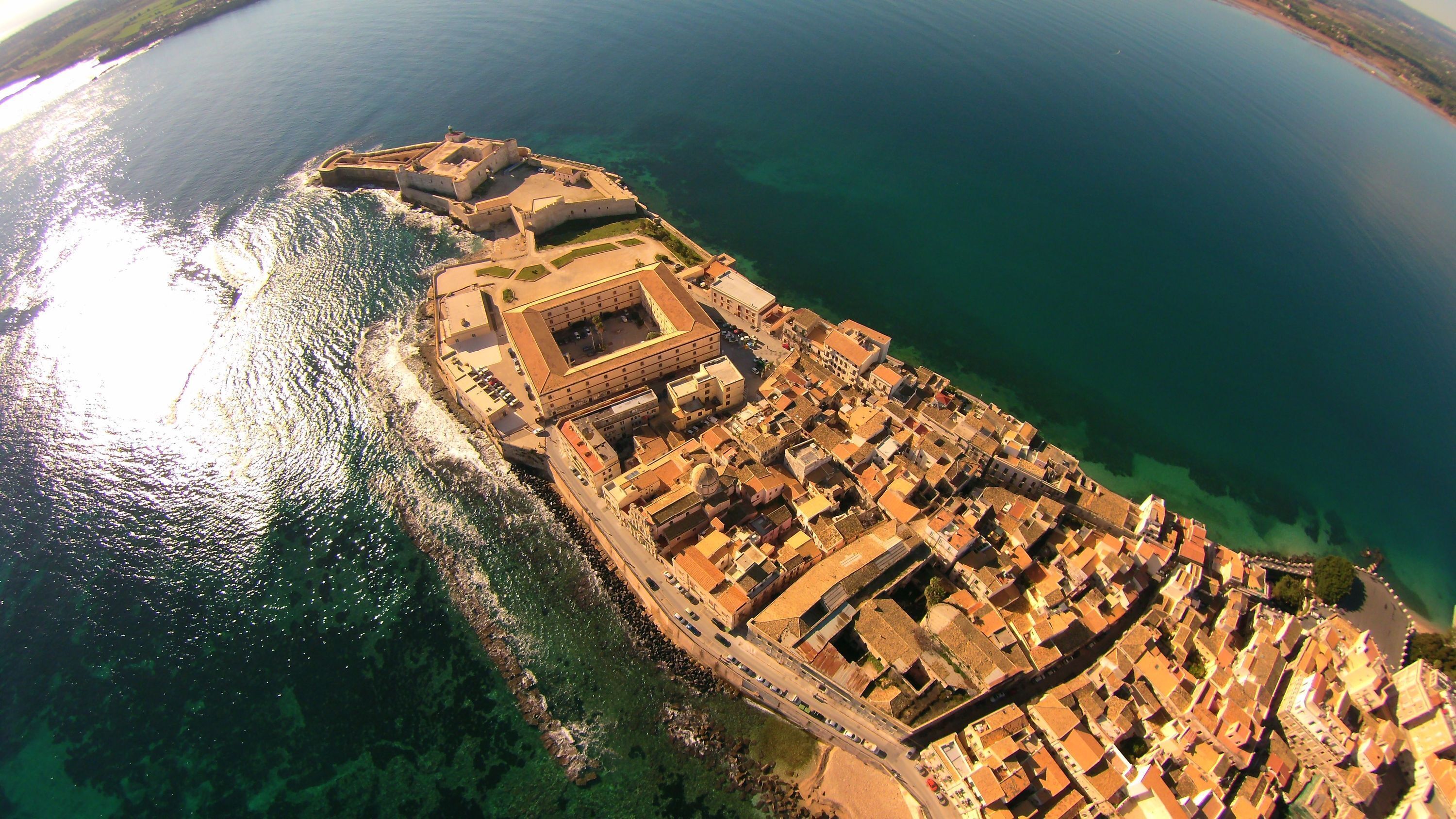 An aerial view of a coastal town with a fortified structure, clear waters, and a dense urban layout.