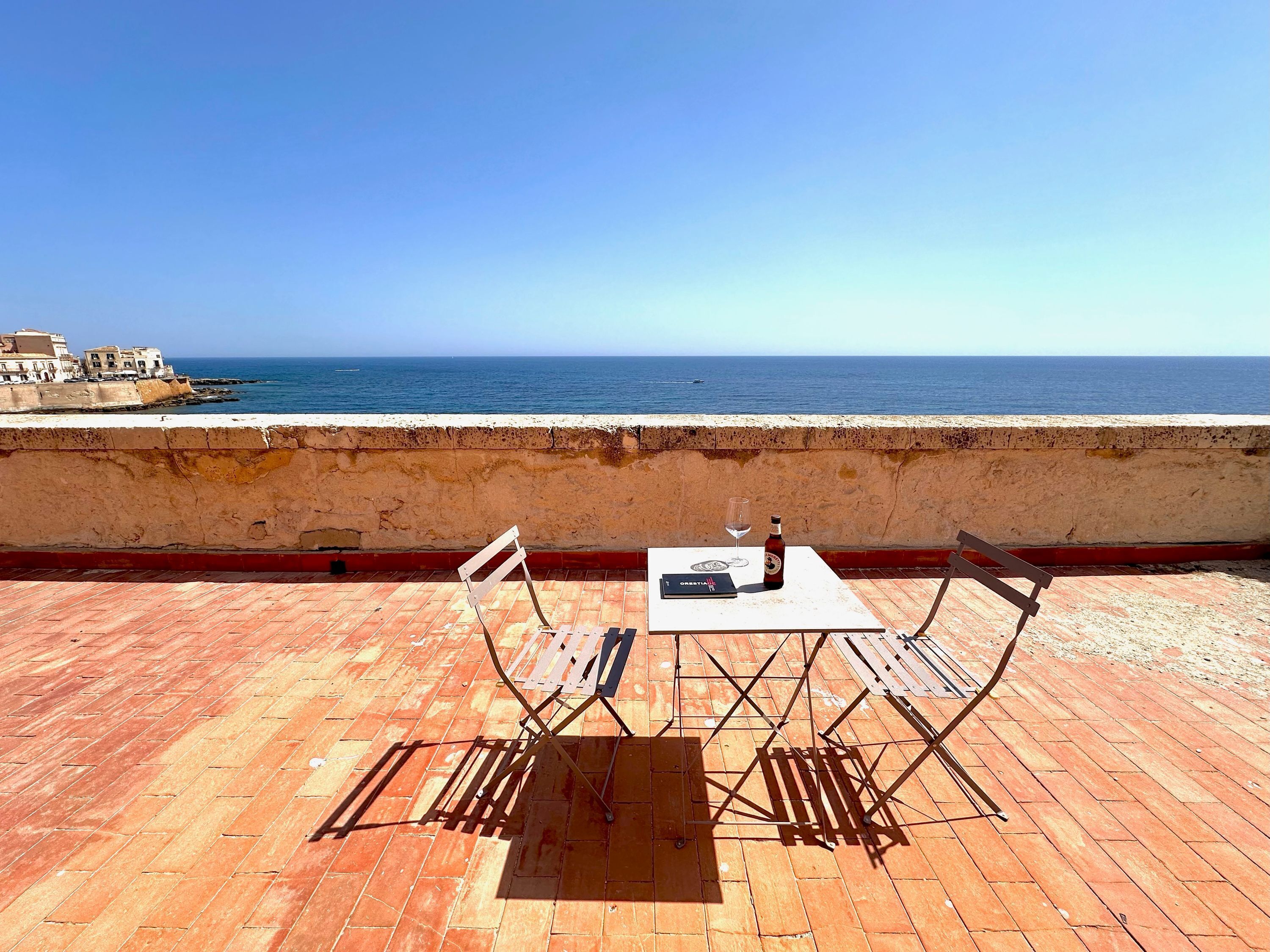 Eine Terrasse mit Tisch und Stühlen, einer Flasche und einem Glas mit Blick auf das Meer.