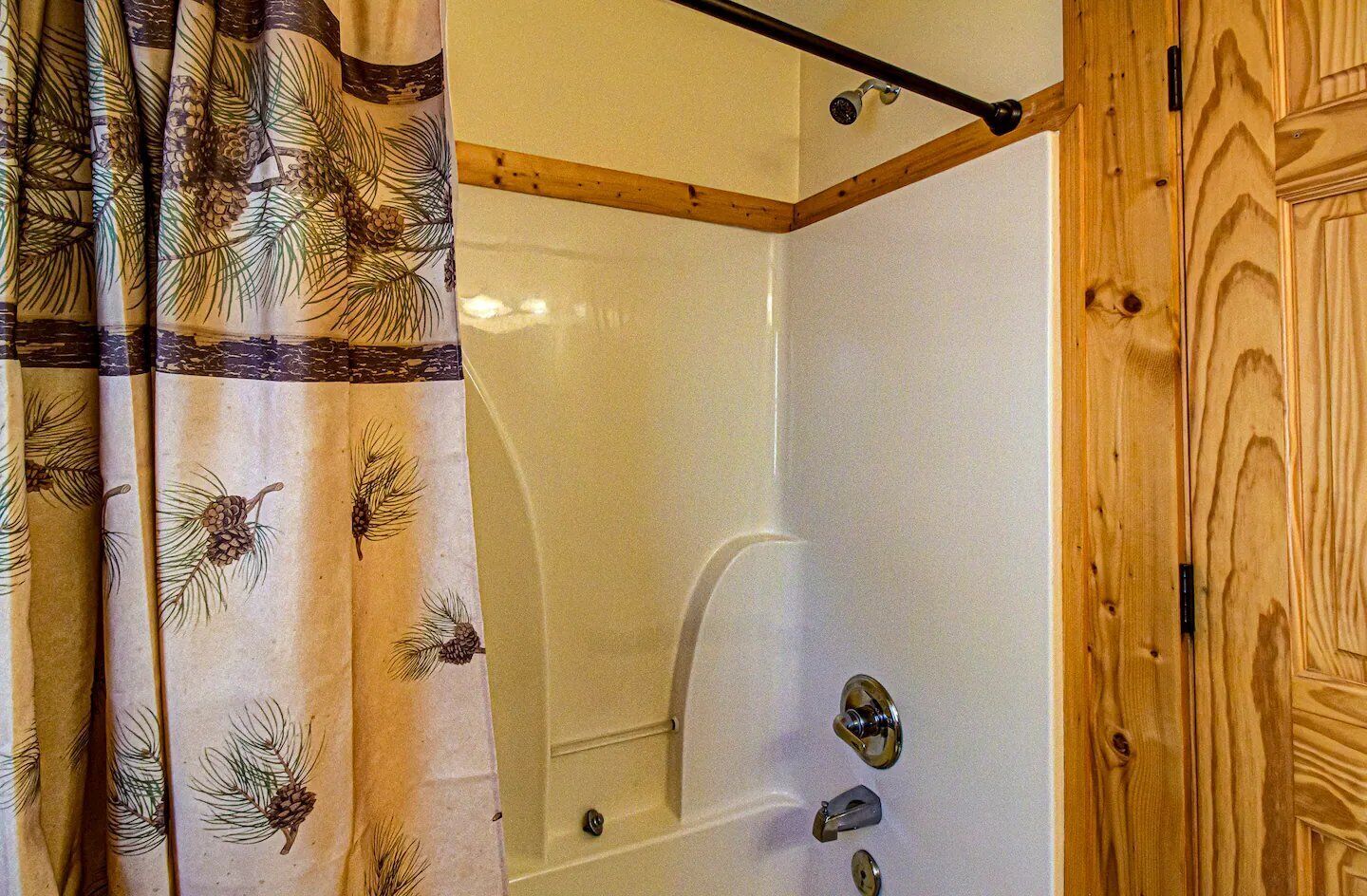 A wooden cabin bathroom with a shower, a wooden door, and a curtain with pine needle patterns.