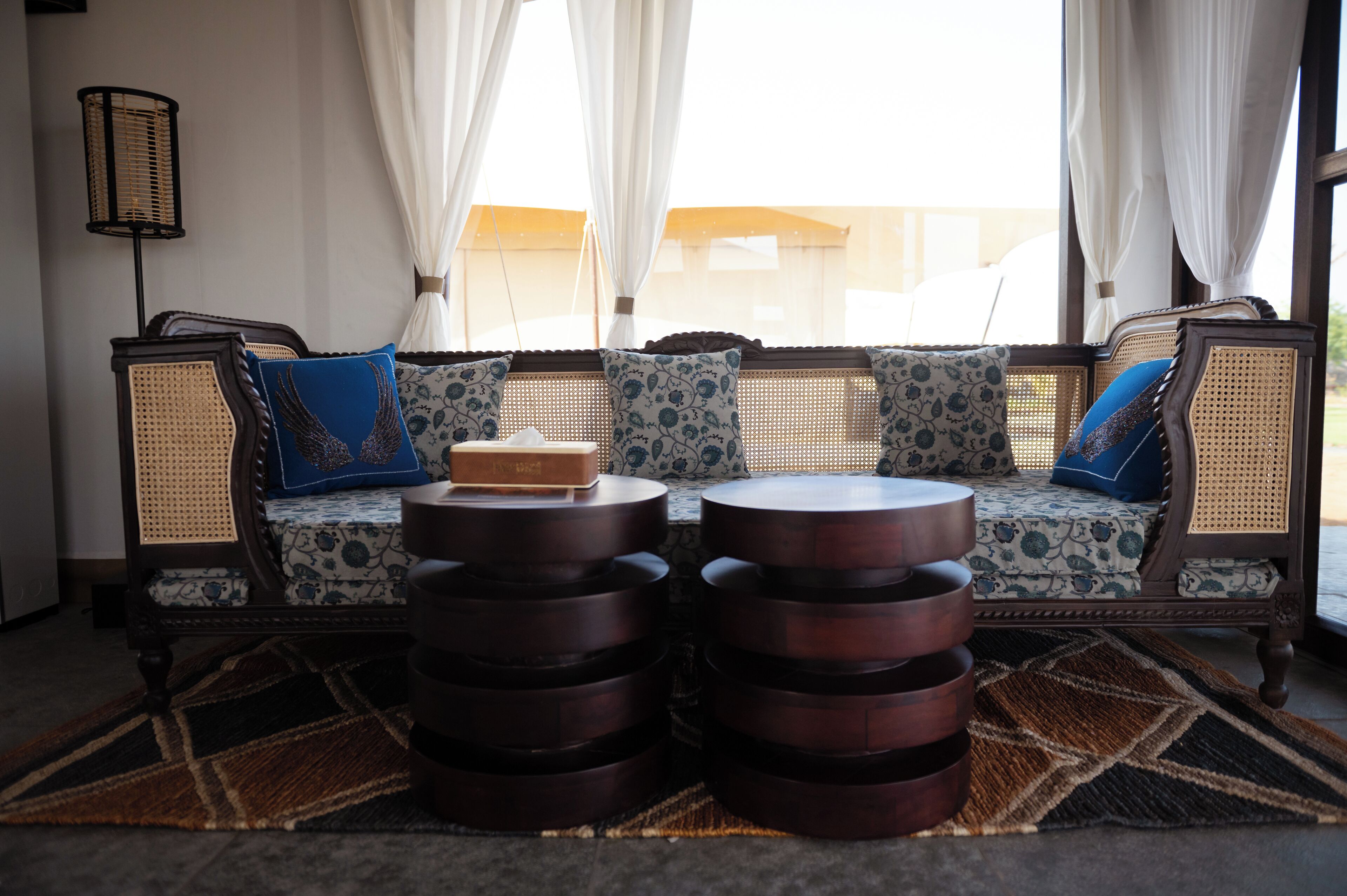 Un sofá de mimbre con cojines estampados, una pequeña mesa de madera y un tapete con diseño en una habitación luminosa con ventanas grandes y cortinas translúcidas.