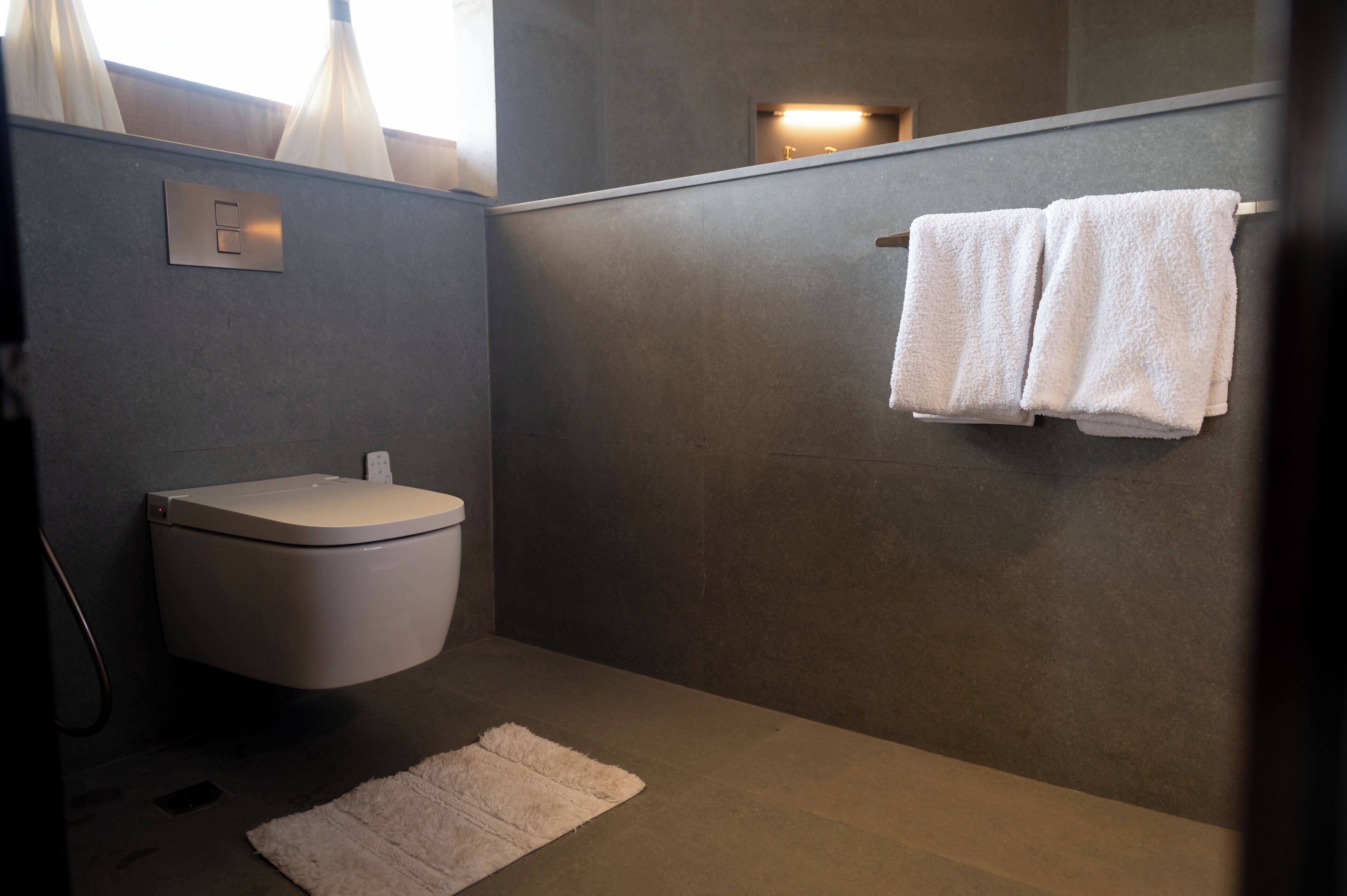 Une salle de bain moderne comprenant des toilettes, un porte-serviettes avec deux serviettes, un luminaire et un petit tapis.