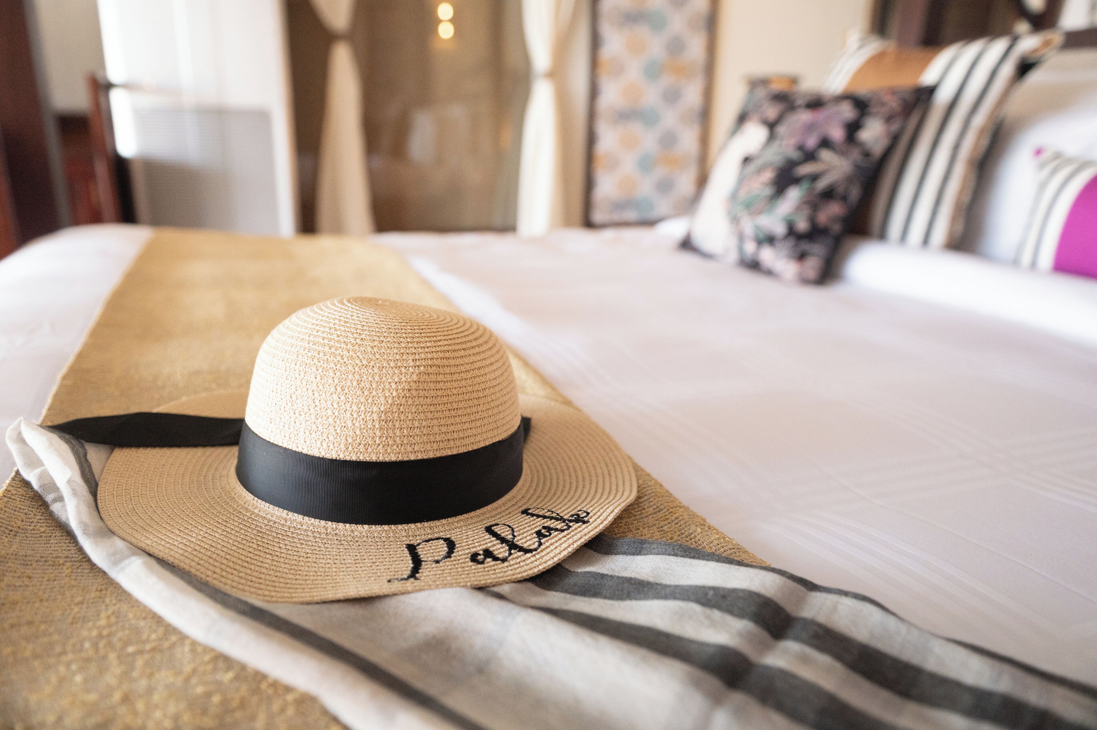 Un sombrero de paja con un lazo negro y la palabra 'Palace' sobre una cama con una manta a rayas.