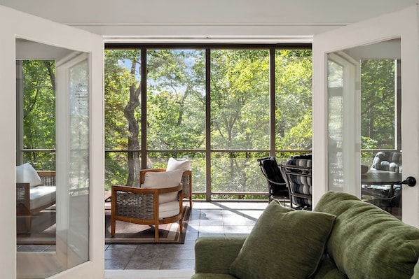 Une pièce dotée de grandes fenêtres, d’un canapé vert, de fauteuils en osier et offrant une vue sur les arbres.
