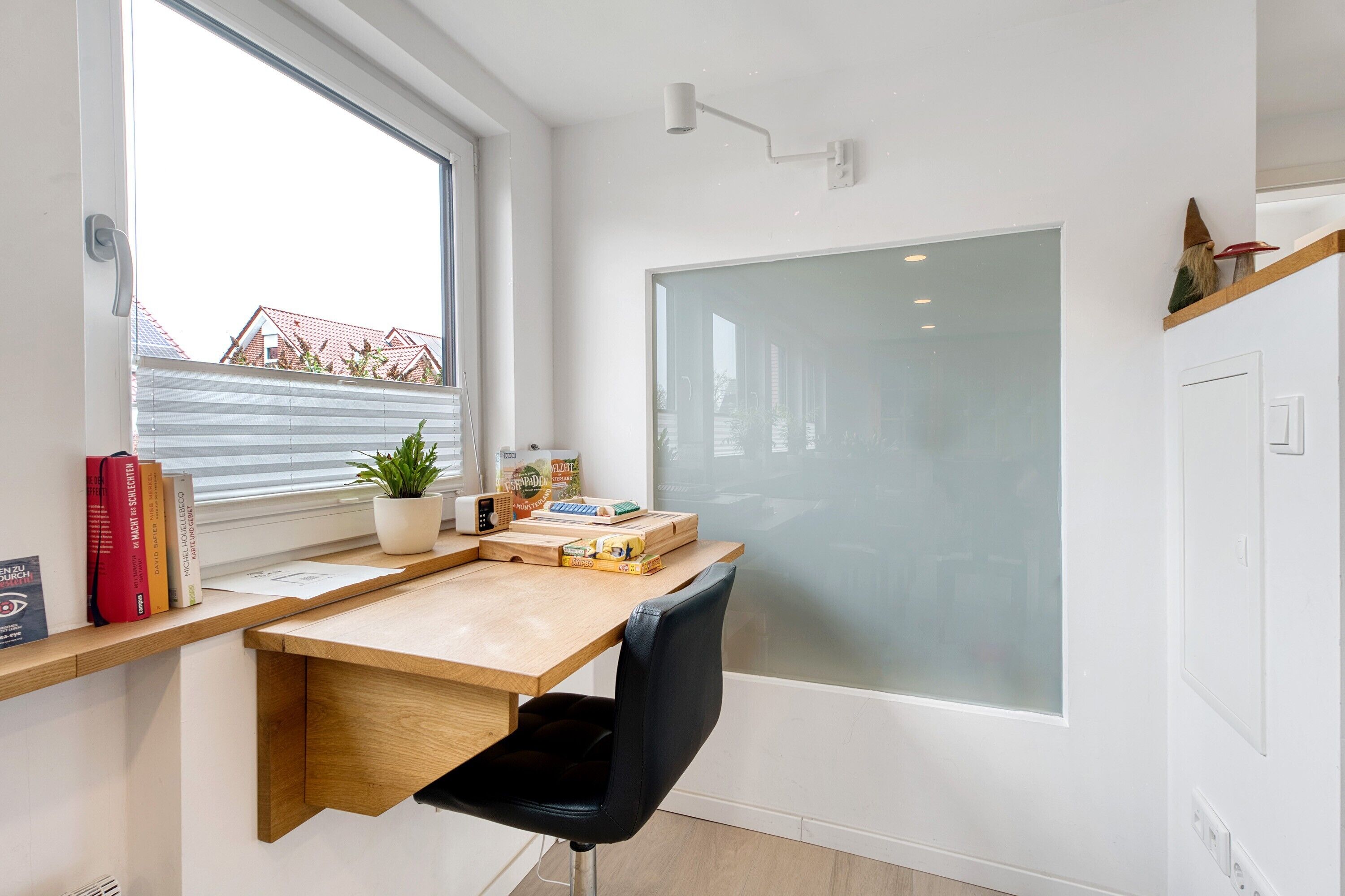 Een compact, modern kantoor met een houten bureau, een zwarte stoel, een raam met zonwering, een potplant en uitzicht op gebouwen.