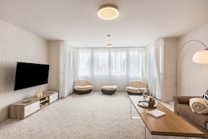Ein modernes Wohnzimmer mit einem Flachbildfernseher, drei beigefarbenen Sofas, einem Holztisch und einem großen Fenster mit durchsichtigen Vorhängen.