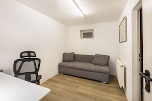 A room with a white table, a black office chair, a grey sofa, a small window, and a radiator.
