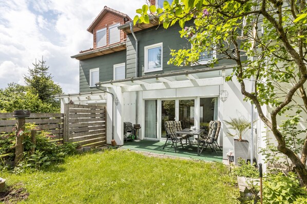 Ein Haus mit Terrasse, Essbereich mit Stühlen, Grill und umzäunter Garten.