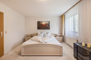 Ein Schlafzimmer mit Bett, Nachttisch, Kommode, einem Gemälde und einem Fenster mit Vorhängen.