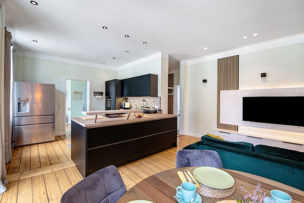 Une cuisine moderne avec un îlot central, un coin repas comprenant une table et des chaises, un canapé et un grand téléviseur fixé au mur.