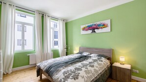 Un dormitorio con cama, mesita de noche, lámpara, ventana con cortinas y un cuadro en la pared.