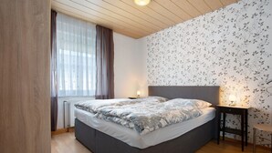 Ein Schlafzimmer mit Bett, Nachttisch, Lampe, Fenster mit Vorhängen und floralem Wandpapier.