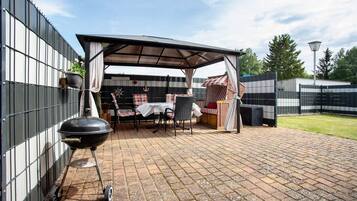 A covered patio area with a barbecue grill, outdoor seating, and a fenced garden.