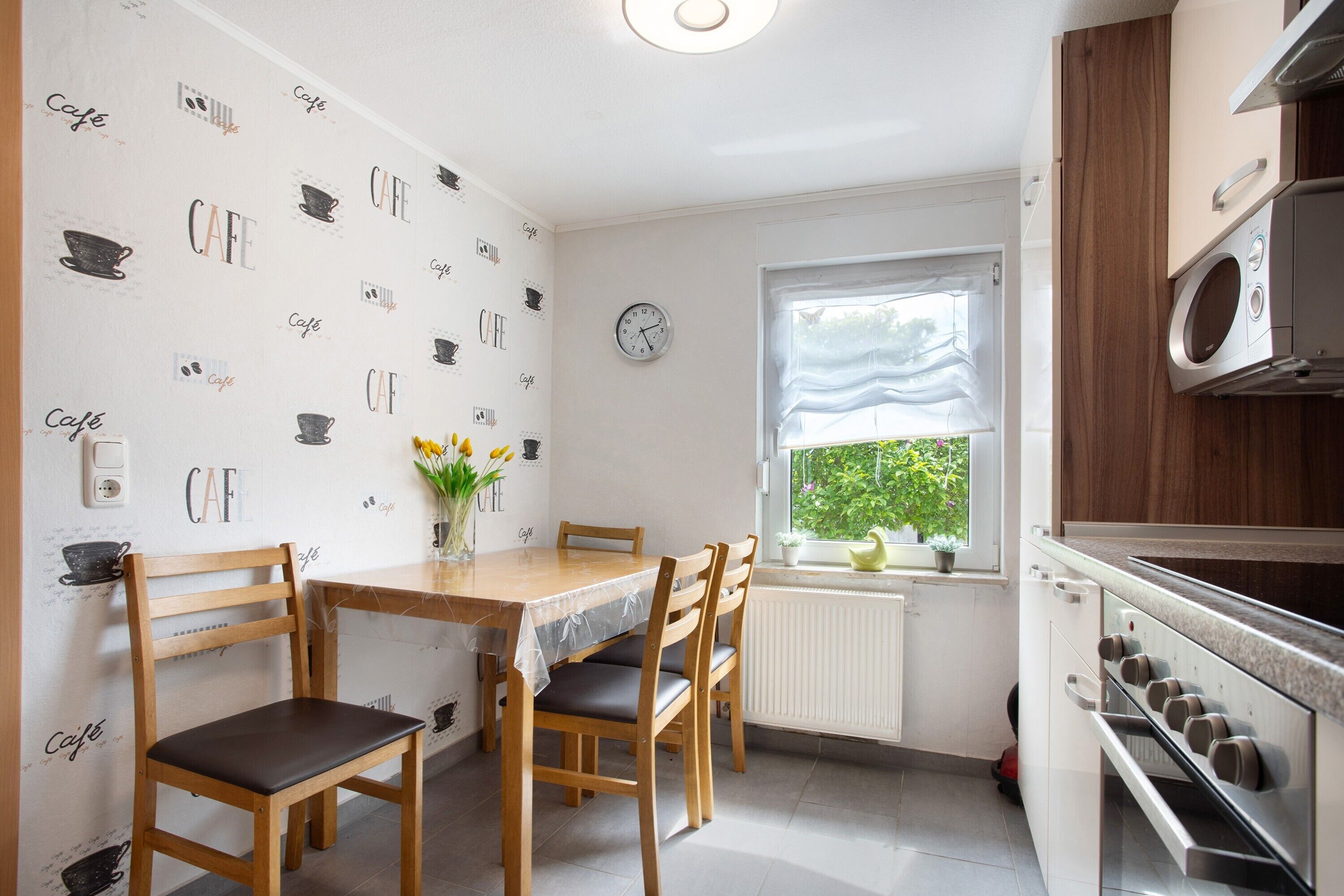 A kitchen with a wooden table, chairs, a clock, and a window with a view of greenery.