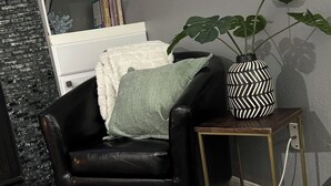 Een gezellig zithoek met een zwarte leren fauteuil, een klein bijzettafeltje met een vaas met dessin, een wit kussen en een plant.