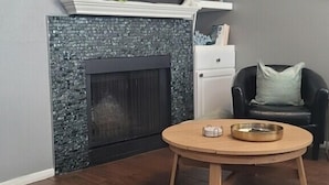 Een woonkamer met een open haard, een flatscreen televisie erboven, een ronde houten salontafel en een zwarte leren fauteuil met kussen.