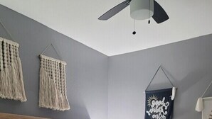 Een slaapkamer met een houten bed, grijze wanden, macramé wanddecoraties, een plafondventilator en een bord aan de muur.