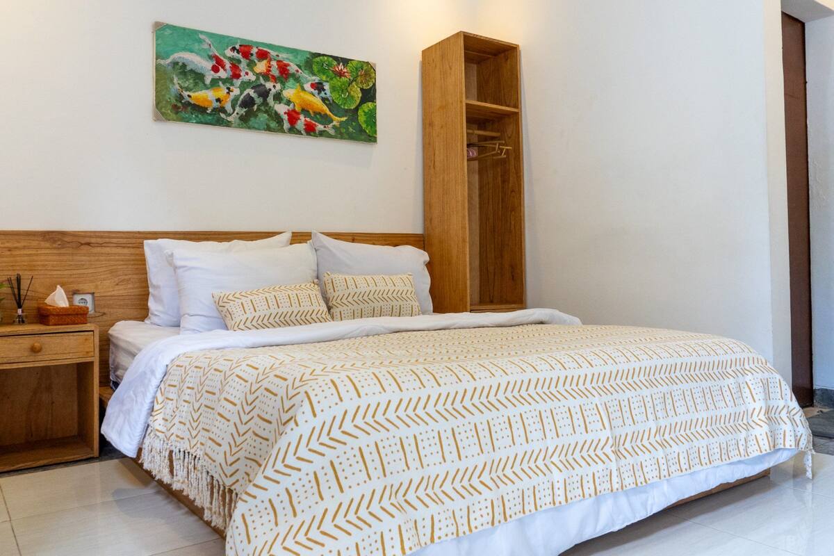 A neatly made bed with patterned bedding, a wooden headboard, a nightstand with a lamp, and a painting of fish on the wall.