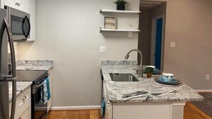 Une cuisine compacte avec des meubles blancs, un four à micro-ondes encastré, un plan de travail en granit, un évier et une petite étagère ornée d’objets décoratifs.