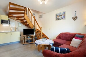 Un salon avec un escalier en bois, un canapé rouge, une petite table recouverte d’une nappe, un téléviseur et un coin cuisine équipé d’un four à micro-ondes.