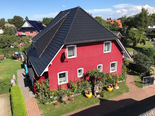 Une maison rouge avec un toit en pente, des panneaux solaires et un jardin agrémenté de fleurs et de chaises.