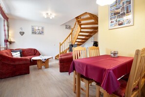 Un salon chaleureux avec un escalier en bois, une table à manger recouverte d’une nappe rouge et un canapé rouge.