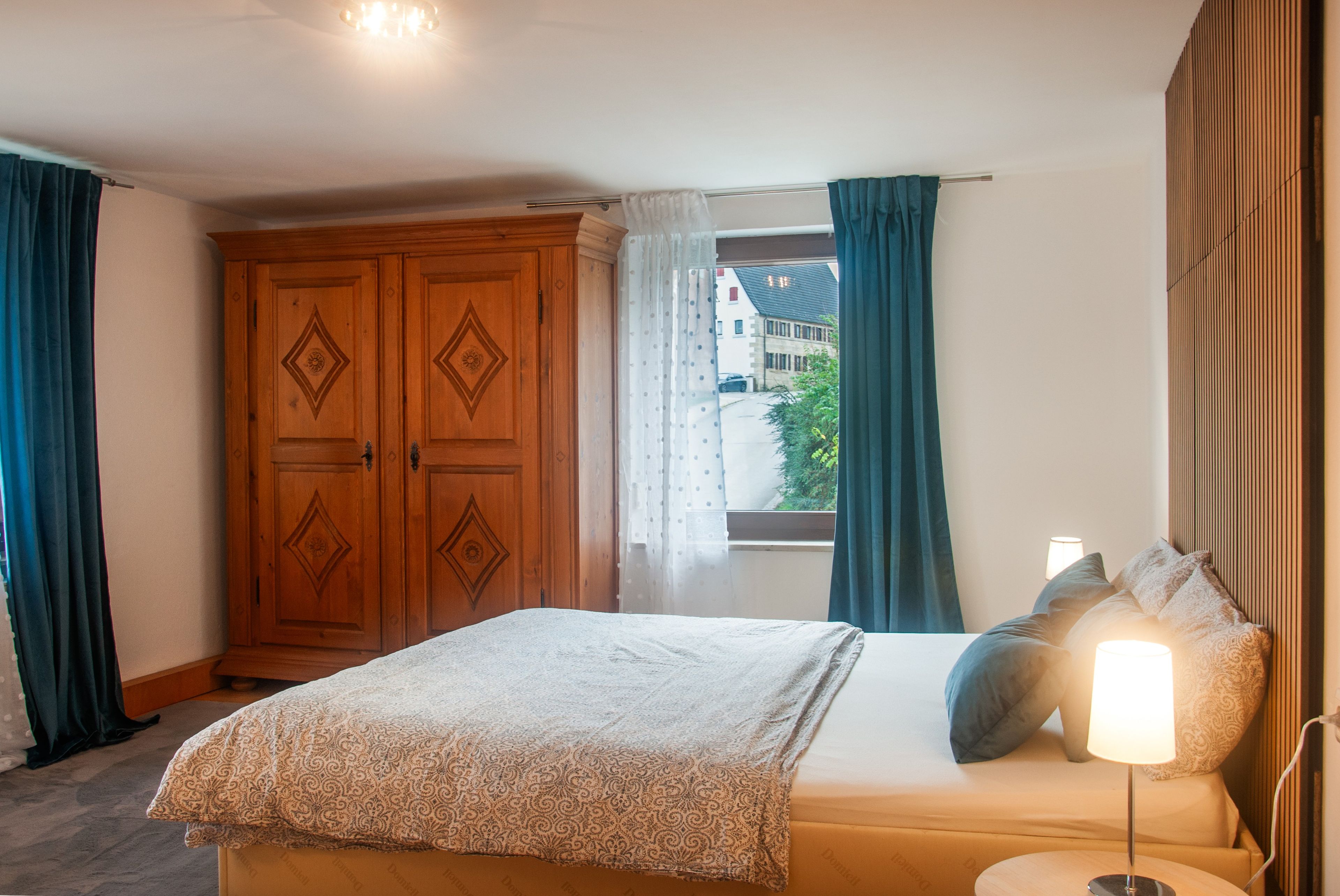 Une chambre à coucher comprenant un lit, une table de chevet, une lampe, une fenêtre avec des rideaux et une armoire en bois.
