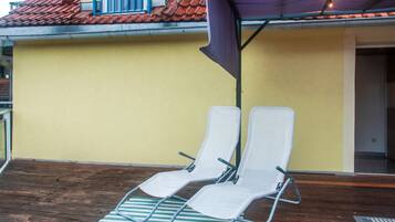 Deux fauteuils blancs sont placés sur une terrasse en bois, agrémentée d’un tapis à rayures, sous un auvent violet, le tout adossé à un mur jaune surmonté d’un toit recouvert de tuiles rouges.