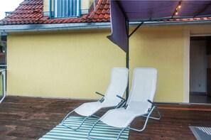 Deux fauteuils blancs sont placés sur une terrasse en bois, agrémentée d’un tapis à rayures, sous un auvent violet, le tout adossé à un mur jaune surmonté d’un toit recouvert de tuiles rouges.