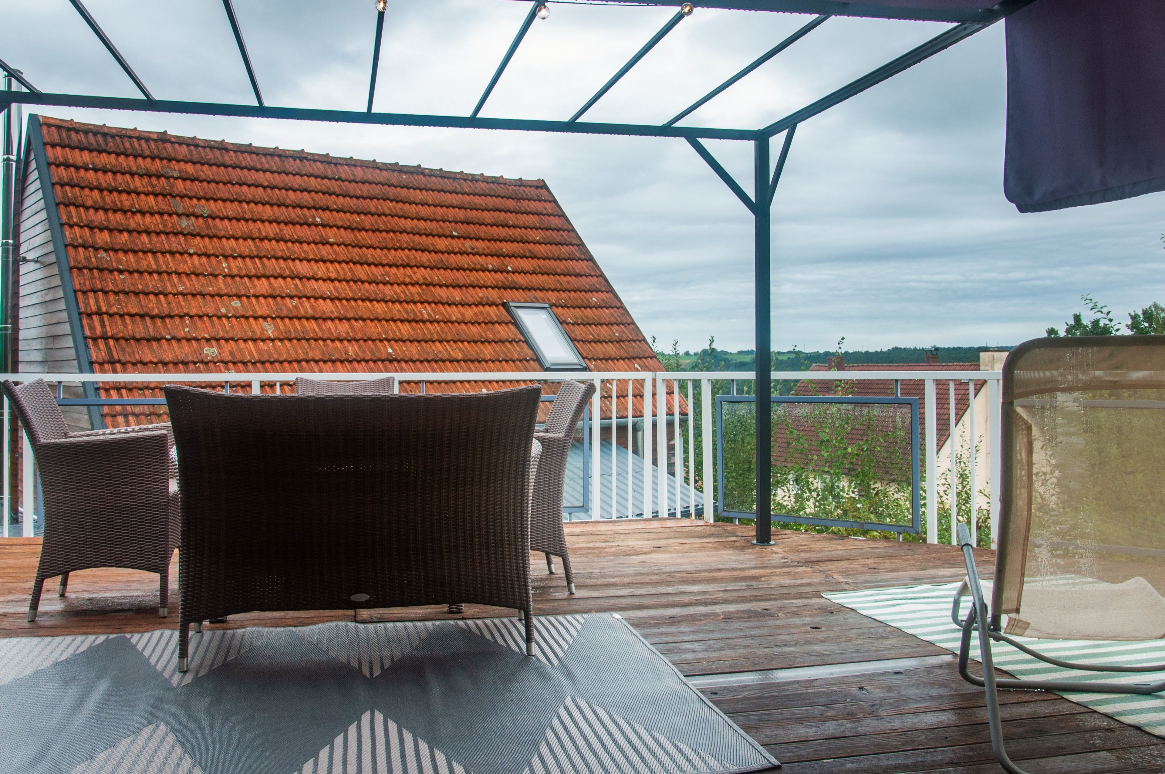 A covered outdoor area with a wooden deck, wicker furniture, and a view of a red-tiled roof building.