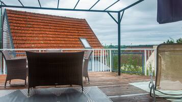 A covered outdoor area with a wooden deck, wicker furniture, and a view of a red-tiled roof building.