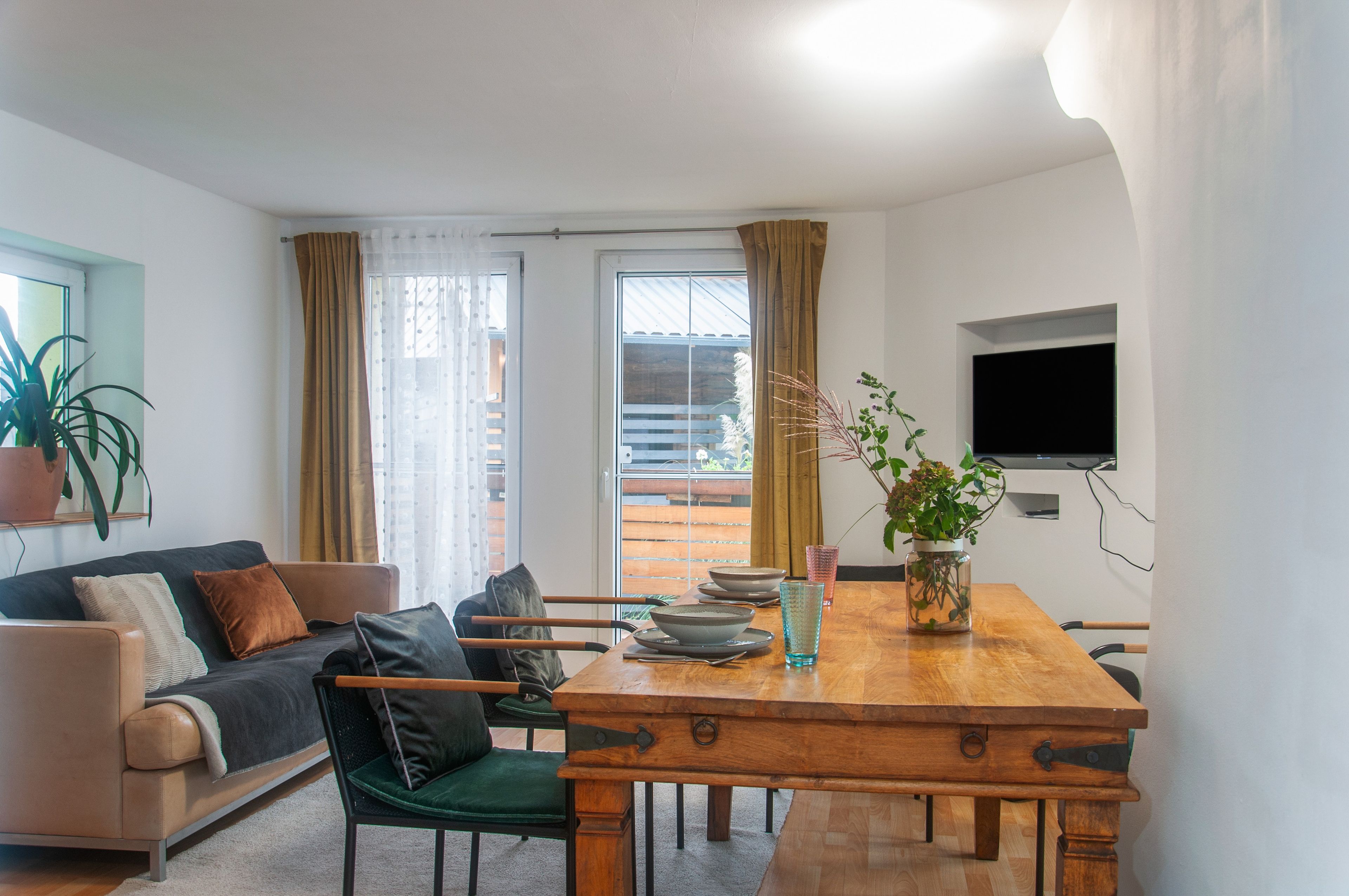 A modern living room with a wooden dining table, a sofa, and a TV mounted on the wall.