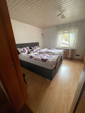 A bedroom with a bed, a window with curtains, a ceiling fan, and a wooden floor.