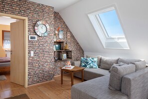A loft-style room with a sloped ceiling, a brick wall, a large window, a sofa, a small table, and a clock.