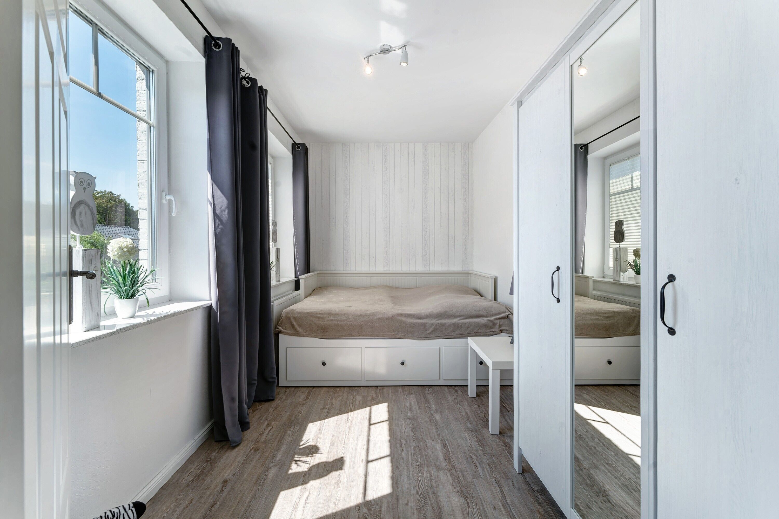 A bedroom with a bed, a small table, a window with curtains, and a view of the outside.